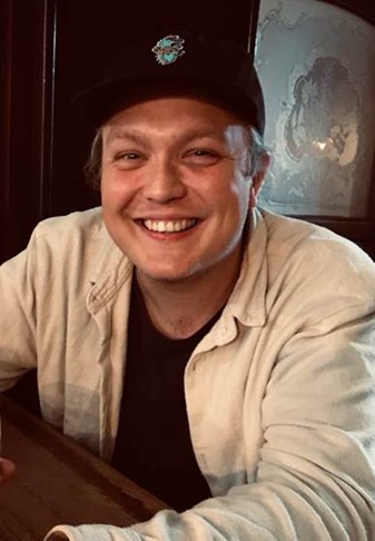 A male academic, smiling, wearing a black baseball cap and wering a cream coloured shirt over a black Tshirt